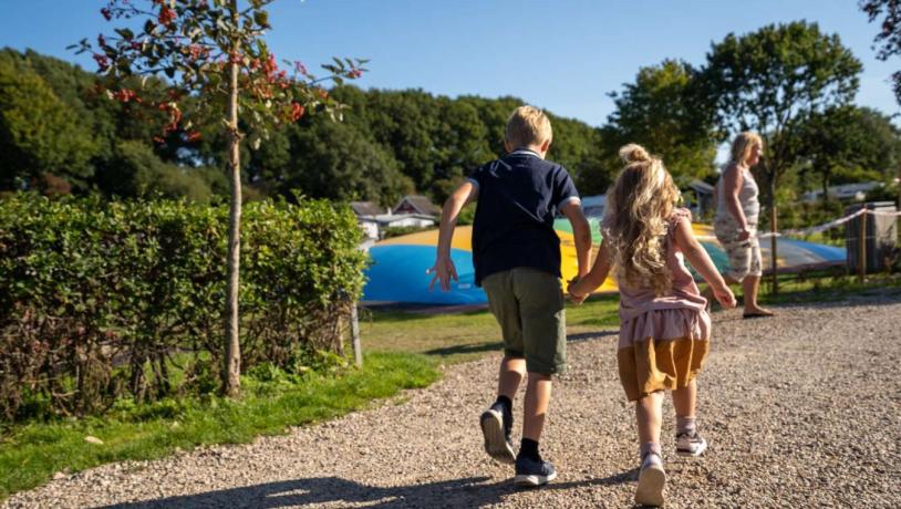 Zwei Kinder halten sich bei vollem Tempo auf dem Weg zum Hüpfkissen auf dem Spielplatz von Horsens City Camping an der Hand – ein Teil der Urlaubsregion Küstenland