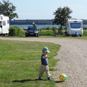 Kleiner Junge spielt mit einem Ball auf dem Rasen von Horsens City Camping mit Wohnwagen und Horsens Fjord im Hintergrund