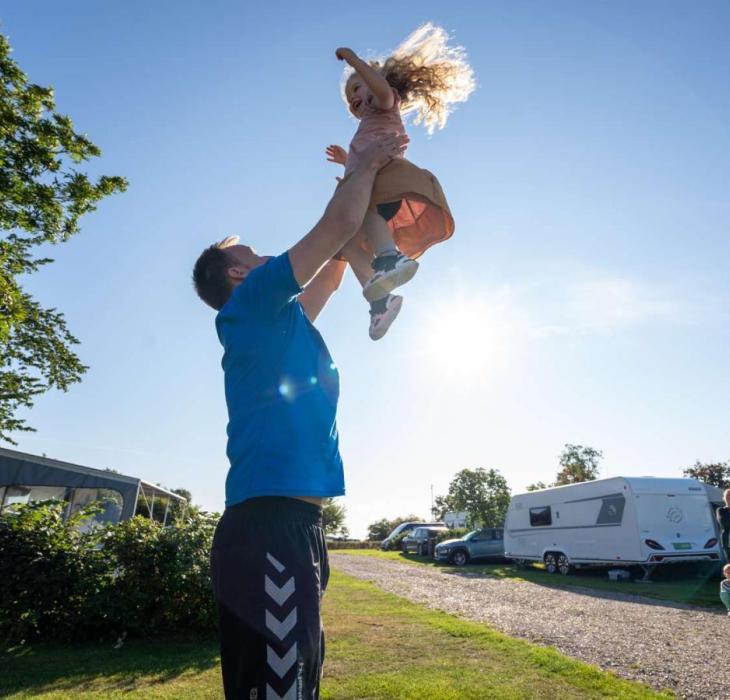 Vater spielt mit seiner Tochter und wirft sie vor dem Wohnwagen bei Horsens City Camping in die Luft – ein Teil der Urlaubsregion Küstenland